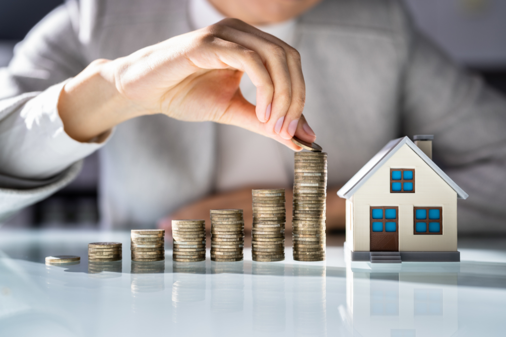 Person stacking coins on a table with a toy-sized house.
