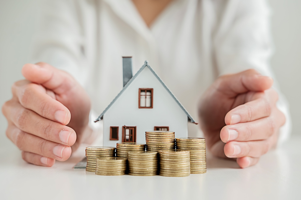 Person's hands on a table surrounding a small house and gold coins.